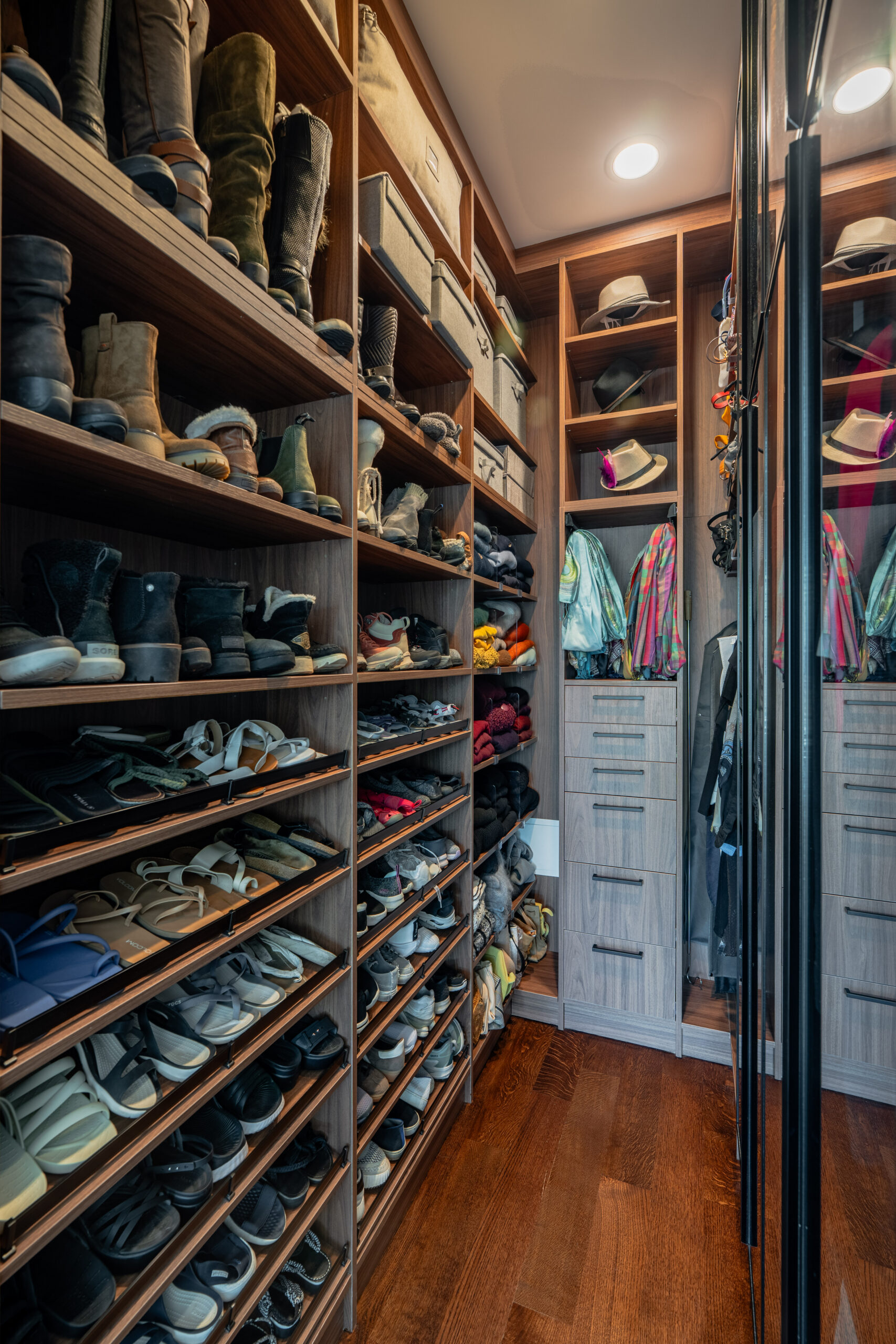 Floor-to-ceiling angled walnut shoe shelving wall with extensive boot and sandal collection, hat cubby display, built-in drawer tower, and colorful hanging garments visible through glass doors in Washington DC luxury walk-in closet