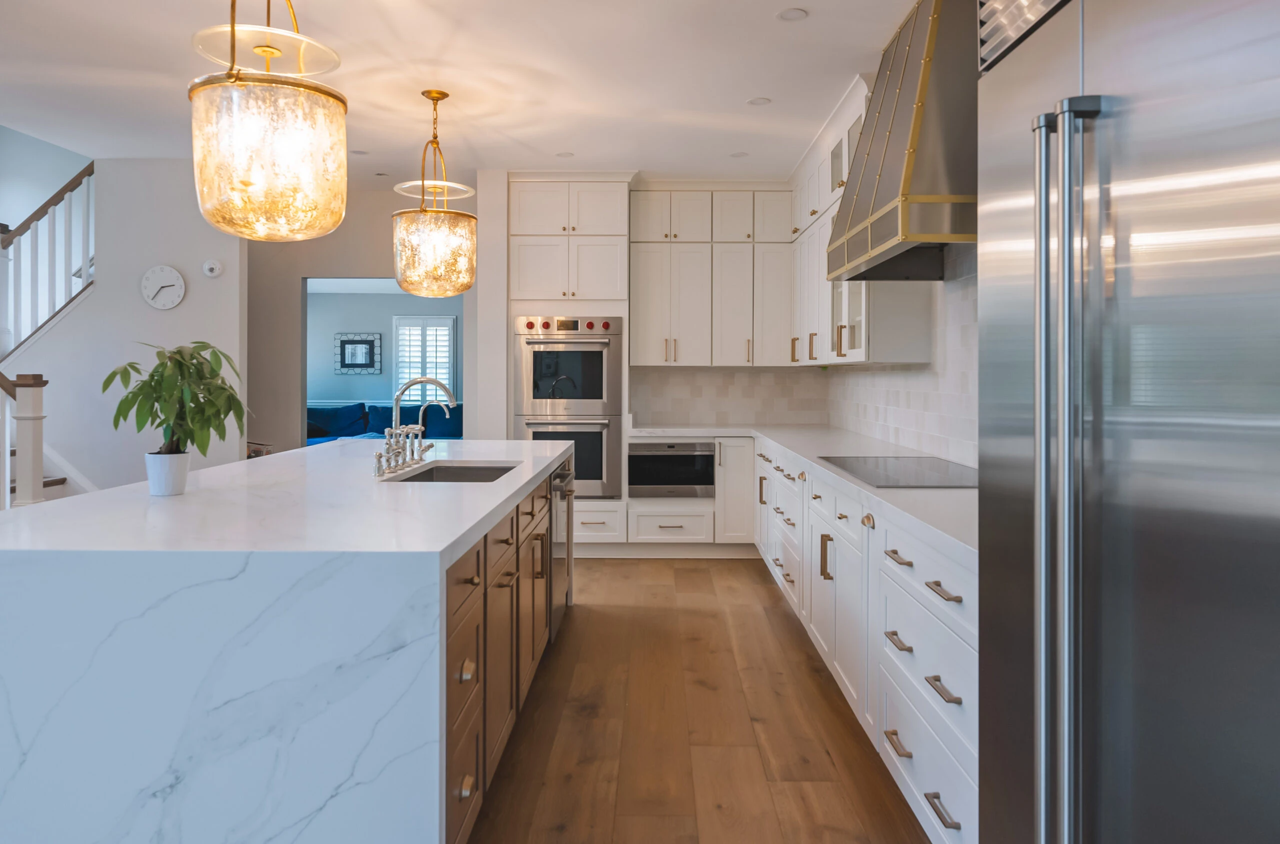 Kitchen remodel in Leesburg, VA featuring floor-to-ceiling white shaker cabinets, large marble quartz island with undermount sink, brass pendant lights, custom range hood with gold trim, and hardwood flooring by Build Design Center