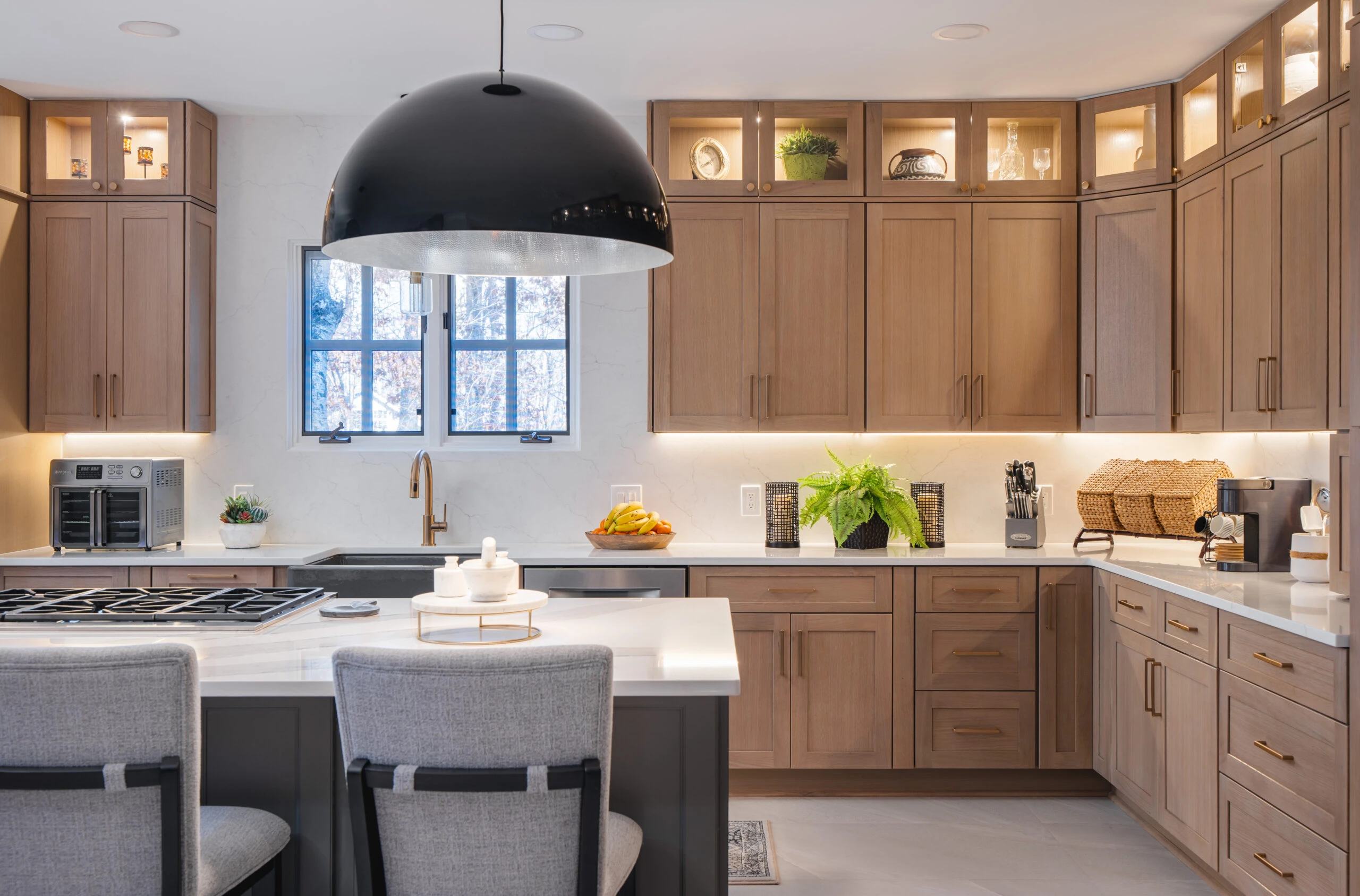 A Cherry Hill, NJ kitchen remodel featuring warm natural wood shaker cabinetry, a large black dome pendant light, quartz countertops, and lit glass-front upper cabinets.