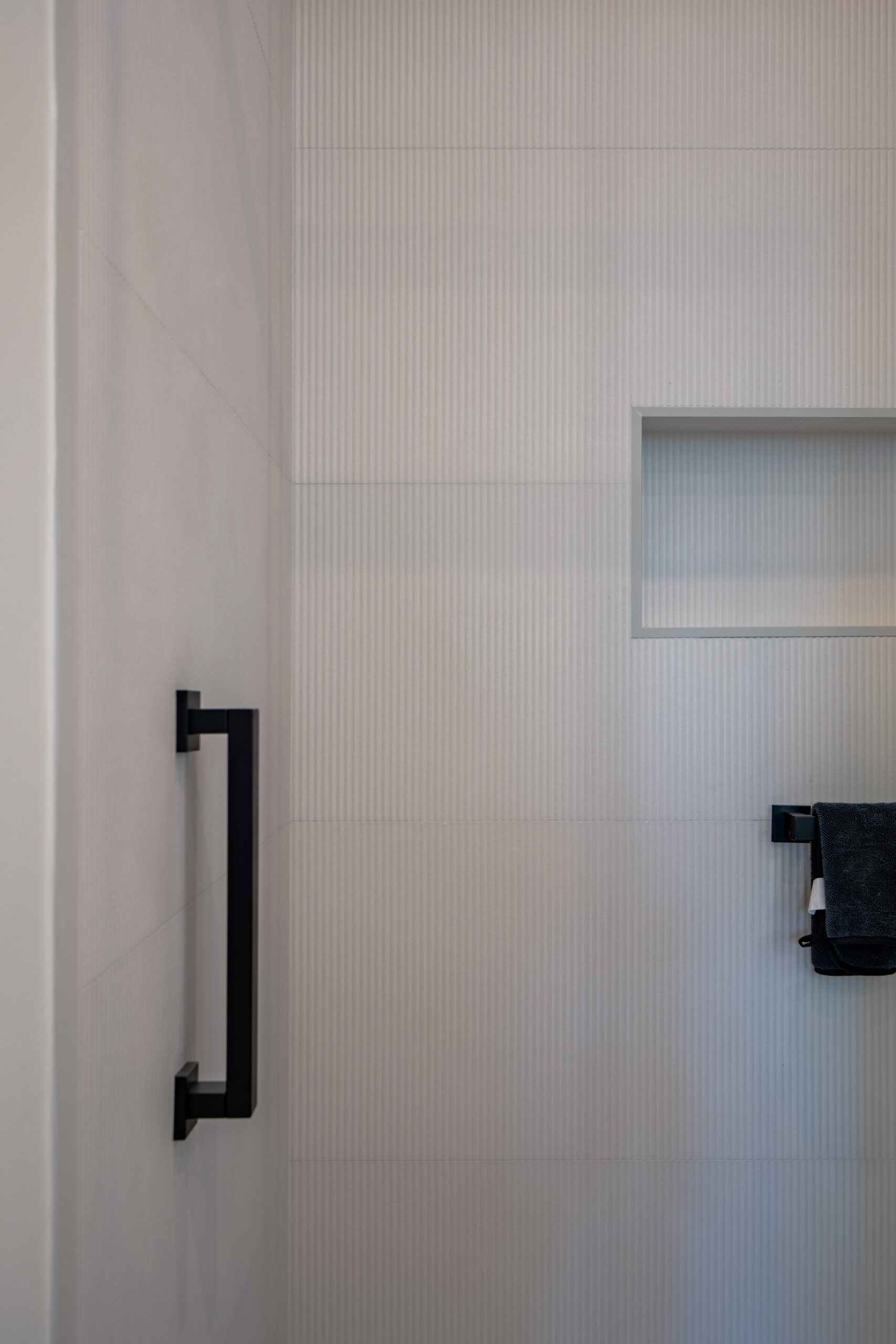 Detail of ribbed white ceramic shower wall tile with recessed built-in shower niche, matte black square door pull handle, and black towel bar in Washington DC luxury bathroom remodel
