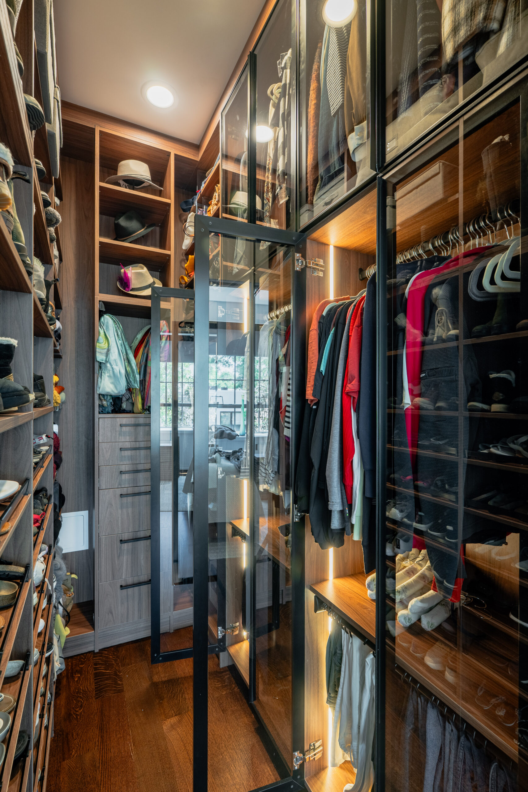 Walk-in closet interior with open black steel framed glass cabinet doors revealing hanging clothes with LED strip lighting, extensive shoe wall shelves, and drawer tower in Washington DC custom cabinetry renovation