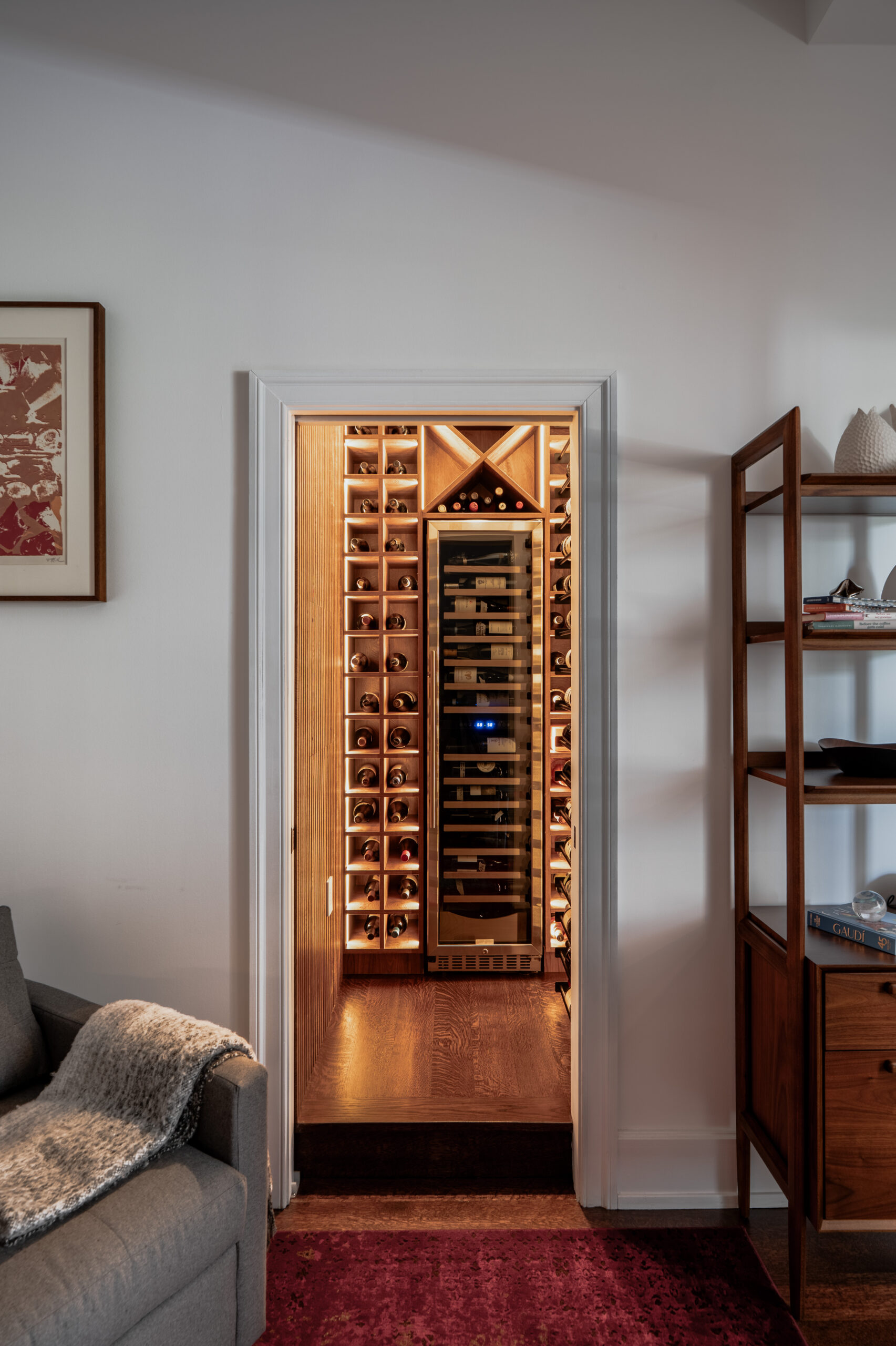 Wine cellar viewed through doorway from living room showing illuminated walnut bottle grid racks and wine refrigerator, with mid-century modern shelving unit and red area rug visible in Washington DC luxury renovation